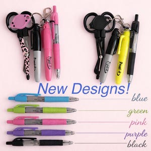 May include: An assortment of pens and keychains are displayed against a light pink background. The pens come in blue, green, pink, purple, and black. The text "New Designs!" is visible.