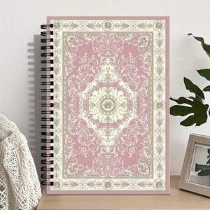 May include: A spiral-bound notebook with a pink and cream-colored ornate rug design on the cover. The rug pattern features floral and scroll motifs. The notebook is displayed on a wooden surface with a white lace garment and a framed picture in the background.