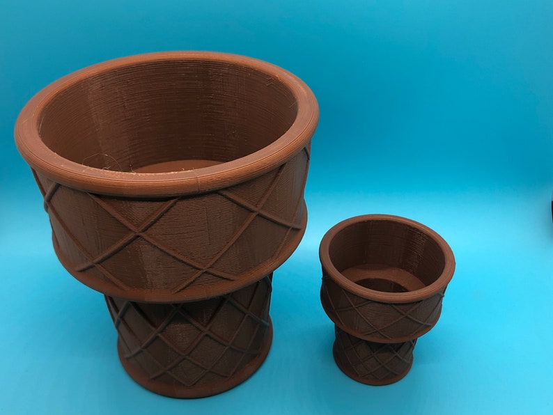 K&ouml;nnte beinhalten: Zwei braune, 3D-gedruckte Pflanzgef&auml;&szlig;e, eines gro&szlig; und eines klein, stehen auf einer blauen Oberfl&auml;che. Die Pflanzgef&auml;&szlig;e haben ein strukturiertes, gewebtes Korbmuster. Das gr&ouml;&szlig;ere Pflanzgef&auml;&szlig; ist etwa 18 cm hoch, das kleinere etwa 8 cm.
