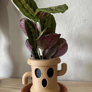 Peut inclure: Un pot en céramique brun en forme de personnage de jeu vidéo avec une expression surprise. Le pot a une petite soucoupe ronde et est rempli d'une plante verte et violette.