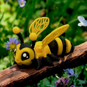 May include: A yellow and black bee toy with large wings and a cute face. The bee is sitting on a brown branch.