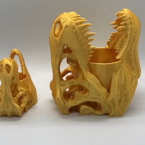 May include: Two gold-colored dinosaur skull planters. The larger planter is open and has a small wooden cup inside. The smaller planter is closed.