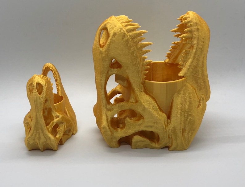 May include: Two gold-colored dinosaur skull planters. The larger planter is open and has a small wooden cup inside. The smaller planter is closed.
