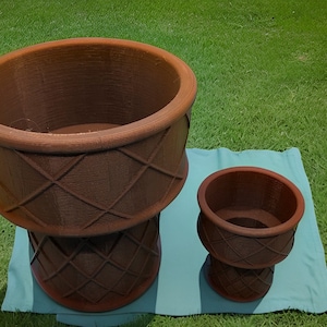 May include: Two brown, decorative planters of different sizes, with a diamond pattern on the base and sides. The planters are sitting on a teal cloth, with a green grass background.