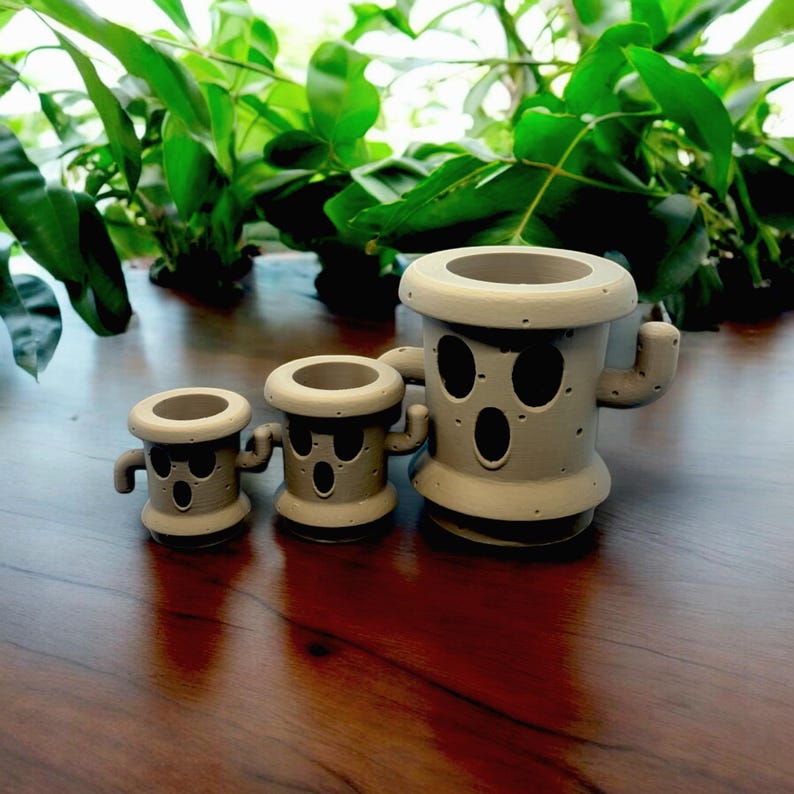 May include: Three gray ceramic planters shaped like cacti with faces. The planters are different sizes and have holes for drainage.