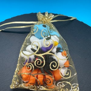 K&ouml;nnte beinhalten: Eine goldene Netz-Tasche gef&uuml;llt mit kleinen, bunten, handgefertigten Polymer Clay-Anh&auml;ngern. Die Anh&auml;nger sind in Form von Geistern, K&uuml;rbissen und Flederm&auml;usen. Die Tasche hat einen goldenen Kordelzug und ist mit einem goldenen Band gebunden.