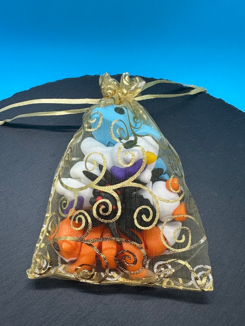 K&ouml;nnte beinhalten: Eine goldene Netz-Tasche gef&uuml;llt mit kleinen, bunten, handgefertigten Polymer Clay-Anh&auml;ngern. Die Anh&auml;nger sind in Form von Geistern, K&uuml;rbissen und Flederm&auml;usen. Die Tasche hat einen goldenen Kordelzug und ist mit einem goldenen Band gebunden.