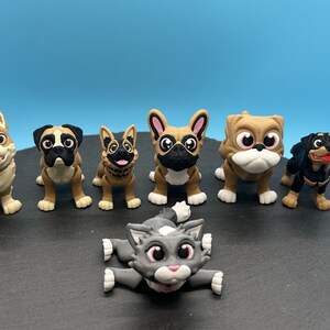May include: A collection of seven small, cartoon-style animal figurines. The figurines are made of a brown, tan, and black plastic. The figurines include a dog, a cat, a bulldog, a rottweiler, a corgi, a french bulldog, and a german shepherd.