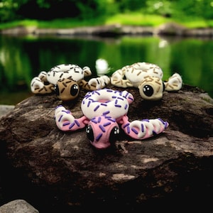 May include: Three small turtle figurines made of polymer clay, each shaped like a donut with different coloured glazes and sprinkles. The turtles are sitting on a rock near a pond.