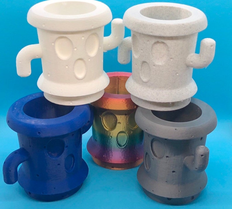 May include: Four 3D printed planters in different colors. Two are white with a textured surface and two are solid colors, one blue and one gray. Each planter has a unique design with two small holes and a handle on the side.