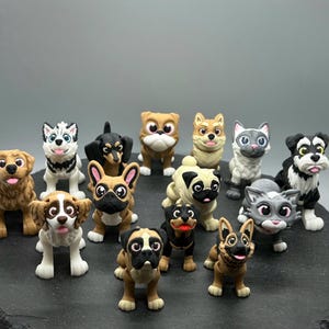 May include: A collection of 13 small, hand-painted figurines of dogs and cats. The figurines are all different breeds and colors, and they are arranged in a circle on a black surface.