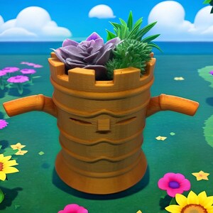 May include: A whimsical, brown, castle-shaped planter with a face and arms, holding a variety of succulents. The planter is filled with a purple rose-like succulent and green spiky plants. The background features a blue sky, water, and colorful flowers.