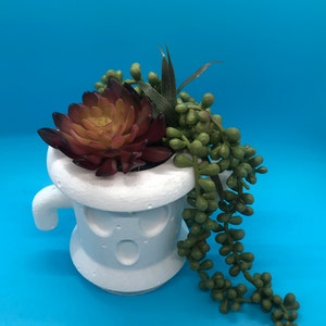 May include: White ceramic ghost-shaped planter with a surprised expression, holding a succulent with burgundy and green leaves and a trailing string of green beads.