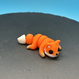 Puede incluir: Figura de zorro naranja hecha a mano con cola y detalles blancos. El zorro tiene ojos negros y patas marrones. La figura está hecha de un material flexible y está tumbada sobre una superficie oscura.