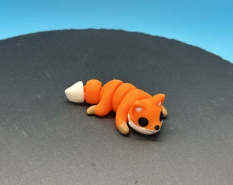 May include: A handcrafted orange fox figurine with a white tail and accents. The fox has black eyes and tan paws. The figurine is made of a pliable material and is lying down on a dark surface.