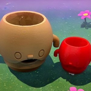 May include: Two whimsical, round planters with cartoon-like faces and small arms. One is tan, the other is red. They sit on a green surface with colorful flowers in the background. These planters are ideal for small plants.