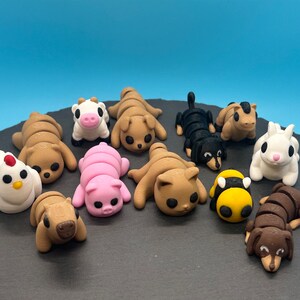 K&ouml;nnte beinhalten: Ein Set aus zehn Fondant-Tierfiguren, darunter eine Kuh, ein Schwein, eine Biene, ein Huhn, eine Katze, ein Hund und ein Kaninchen. Die Figuren sind alle in einer zusammengerollten Position und sollen schlafend aussehen. Die Figuren befinden sich auf einer schwarzen Oberfl&auml;che.