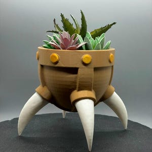 May include: A brown and white 3D printed planter with three legs shaped like horns. The planter is filled with green and pink succulents.