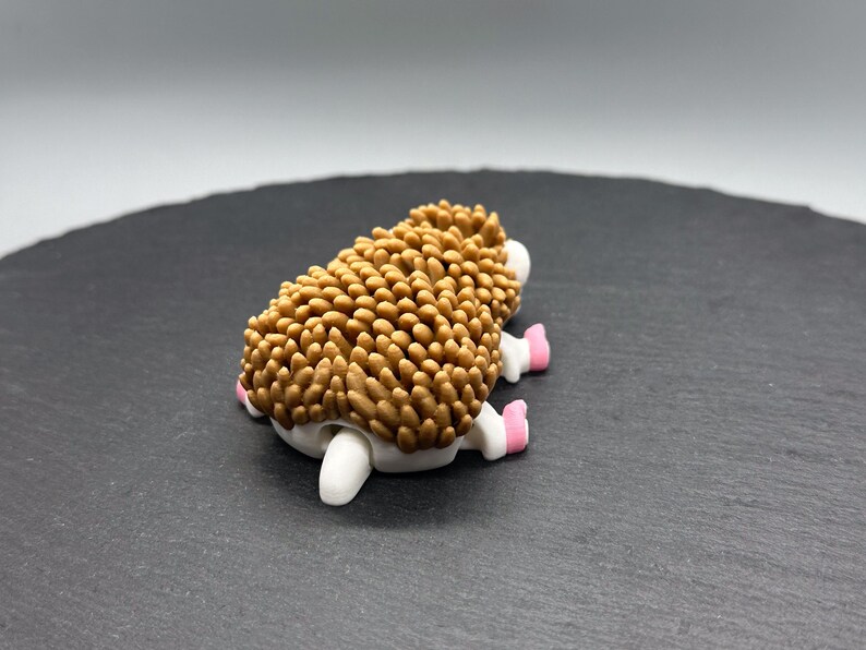Puede incluir: Figurita artesanal de erizo, con un dorso marr&oacute;n texturizado, vientre blanco y patas rosas y blancas. La figurita est&aacute; sobre una superficie gris oscuro.