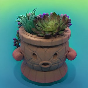 Puede incluir: Una maceta de color terracota con un diseño de cara de dibujos animados, que contiene una variedad de suculentas artificiales. La maceta tiene una base redonda y dos asas pequeñas y redondeadas. Las suculentas son verdes, moradas y verde grisáceo.