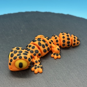 May include: Orange and black segmented toy gecko with a large yellow eye. The gecko is posed on a black surface.