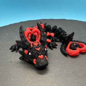 Op de afbeelding: Een zwart en rood 3D-geprint drakenbeeldje met hartvormige details. De draak ligt op zijn buik met zijn kop naar de zijkant gedraaid.