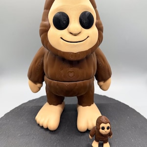 May include: A brown Bigfoot figurine with a light tan face and feet, and black eyes. A smaller version of the figurine is also present. The figures are on a dark grey surface.