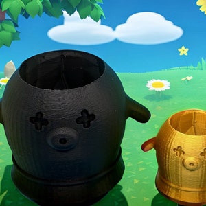 May include: Two 3D-printed planters, one black and one gold, each with a round body, small arms, and a face with cross-shaped eyes and a circular nose. The planters are set against a cartoon background of a green field and blue sky.