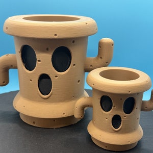May include: Two brown ceramic planters shaped like cacti with black eyes and mouths. The larger planter has two arms and the smaller planter has one arm.