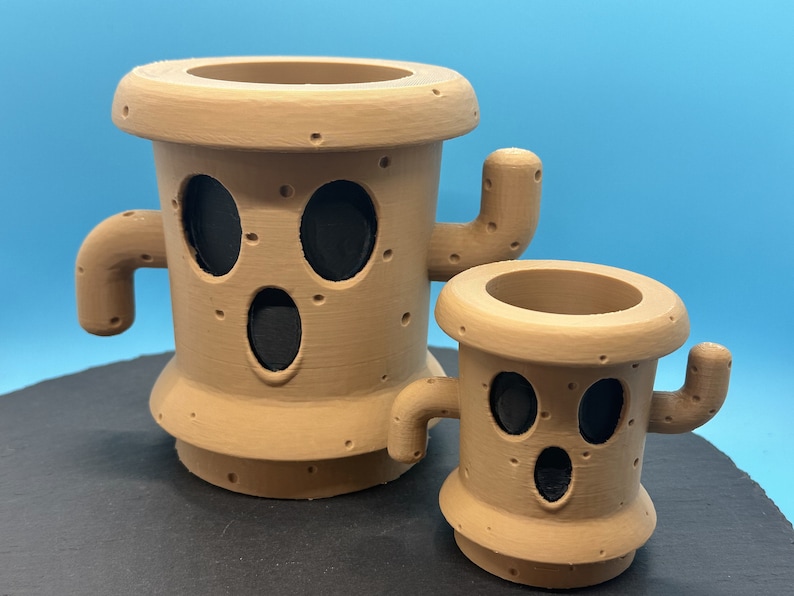 May include: Two brown ceramic planters shaped like cacti with black eyes and mouths. The larger planter has two arms and the smaller planter has one arm.