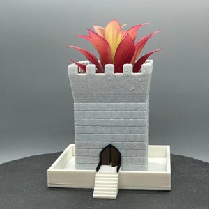 May include: White ceramic planter shaped like a castle tower with a red and yellow bromeliad plant growing inside. The planter has a small door and steps leading up to it.