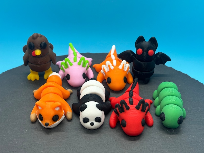 K&ouml;nnte beinhalten: Eine Sammlung von acht kleinen, bunten, 3D-gedruckten Tierfiguren. Die Figuren umfassen einen Truthahn, einen Fuchs, einen Panda, eine Raupe, einen roten Drachen, einen orangefarbenen Drachen, einen rosa Drachen und eine schwarze Fledermaus.