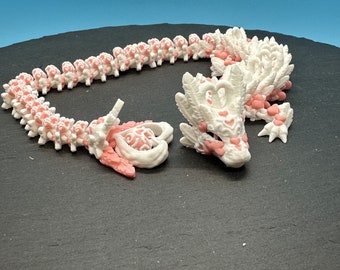 May include: A white and pink 3D printed dragon necklace with a heart design. The dragon is curled up in a circle with its tail in its mouth.