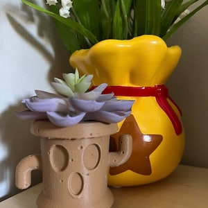 May include: A small brown planter shaped like a tree stump with a purple succulent plant inside. The planter has a brown star on the front and a brown arm on each side. The planter is sitting in front of a yellow ceramic pot shaped like a money bag with a red ribbon around the top.