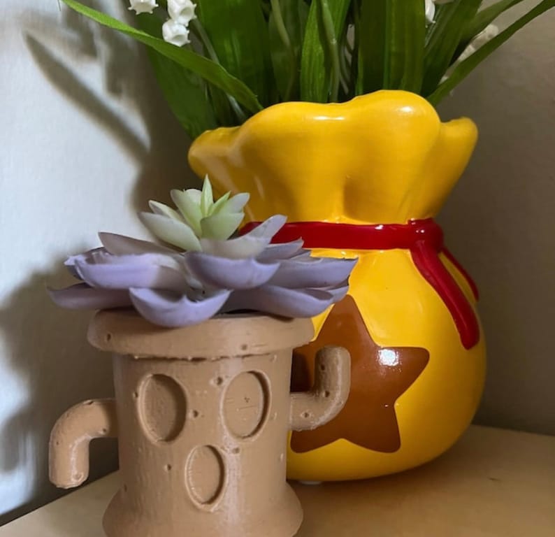 May include: A small brown planter shaped like a tree stump with a purple succulent plant inside. The planter has a brown star on the front and a brown arm on each side. The planter is sitting in front of a yellow ceramic pot shaped like a money bag with a red ribbon around the top.
