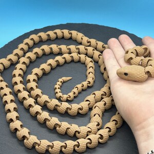 May include: A brown snake toy made of interlocking segments. The snake is coiled on a black surface and a hand is holding the head of the snake.