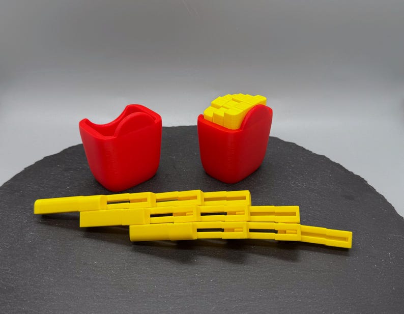 May include: A set of 3D-printed toys resembling fast food items. The set includes a red container, a container of fries, and a yellow, articulated toy. The fries are yellow and the containers are red.