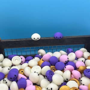 May include: A wooden crate filled with a variety of colorful, small, 3D printed figurines. The figurines are shaped like round, flat, cartoon faces with different expressions. The figurines are in shades of white, purple, pink, gold, and brown.