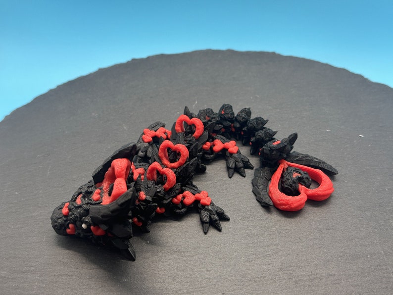 Op de afbeelding: Een zwart en rood 3D-geprint drakenbeeldje met hartvormige details. De draak ligt op zijn zij met zijn kop naar de zijkant gedraaid.