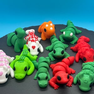 K&ouml;nnte beinhalten: Eine Sammlung von zehn kleinen, bunten Fondantfiguren in Form von Eidechsen und Drachen. Die Figuren sind auf einer schwarzen Schieferfl&auml;che angeordnet.
