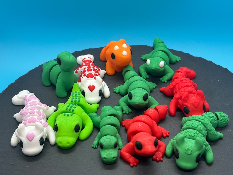 K&ouml;nnte beinhalten: Eine Sammlung von zehn kleinen, bunten Fondantfiguren in Form von Eidechsen und Drachen. Die Figuren sind auf einer schwarzen Schieferfl&auml;che angeordnet.