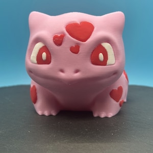May include: A pink 3D printed figurine of a Pokemon character with red hearts on its head and body. The figurine has white eyes with red pupils.