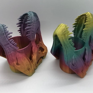 May include: Two rainbow-colored dinosaur skull-shaped pencil holders. The holders are made of 3D printed plastic and have a cylindrical opening in the center.