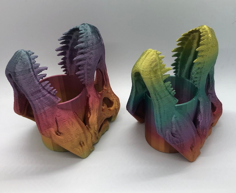 May include: Two rainbow-colored dinosaur skull-shaped pencil holders. The holders are made of 3D printed plastic and have a cylindrical opening in the center.