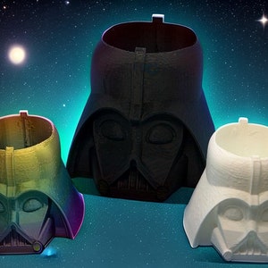 Op de afbeelding: Drie Darth Vader helmvormige plantenbakken in zwart, wit en regenboogkleuren. De plantenbakken staan tegen een sterrenhemel achtergrond. De plantenbakken zijn ontworpen met de iconische helmkenmerken.