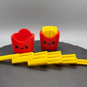 May include: A red container and a red container filled with yellow rectangular shapes, resembling french fries. The containers have smiling faces. Several yellow rectangular shapes are arranged in front of the containers.