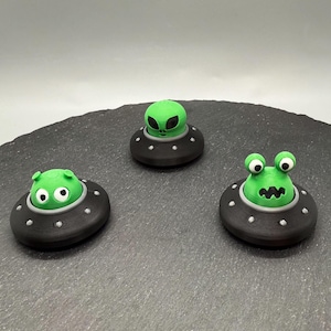 May include: Three whimsical, handmade alien figurines in black UFOs with green alien heads. Each alien has a unique expression, with one featuring large eyes, another with a simple face, and the third with a wide, toothy grin. The UFOs have white dot details.