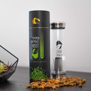 May include: A black and green glass water infuser bottle with a stainless steel lid and a black and gold cardboard tube. The tube has the text "mayj ECO BRAND" and "Enjoy earth in a luxurious way" printed on it. The bottle is sitting on a dark brown surface with dried flowers scattered around it.