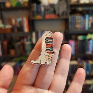 May include: A clear acrylic pin featuring a white cartoon dinosaur holding a stack of five colorful books. The books are stacked vertically in shades of red, blue, and pink. The dinosaur has a simple outline, a small smile, and a curved tail.
