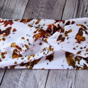 May include: White headband with a brown and gold cow print pattern. The headband is twisted and has a knotted design.
